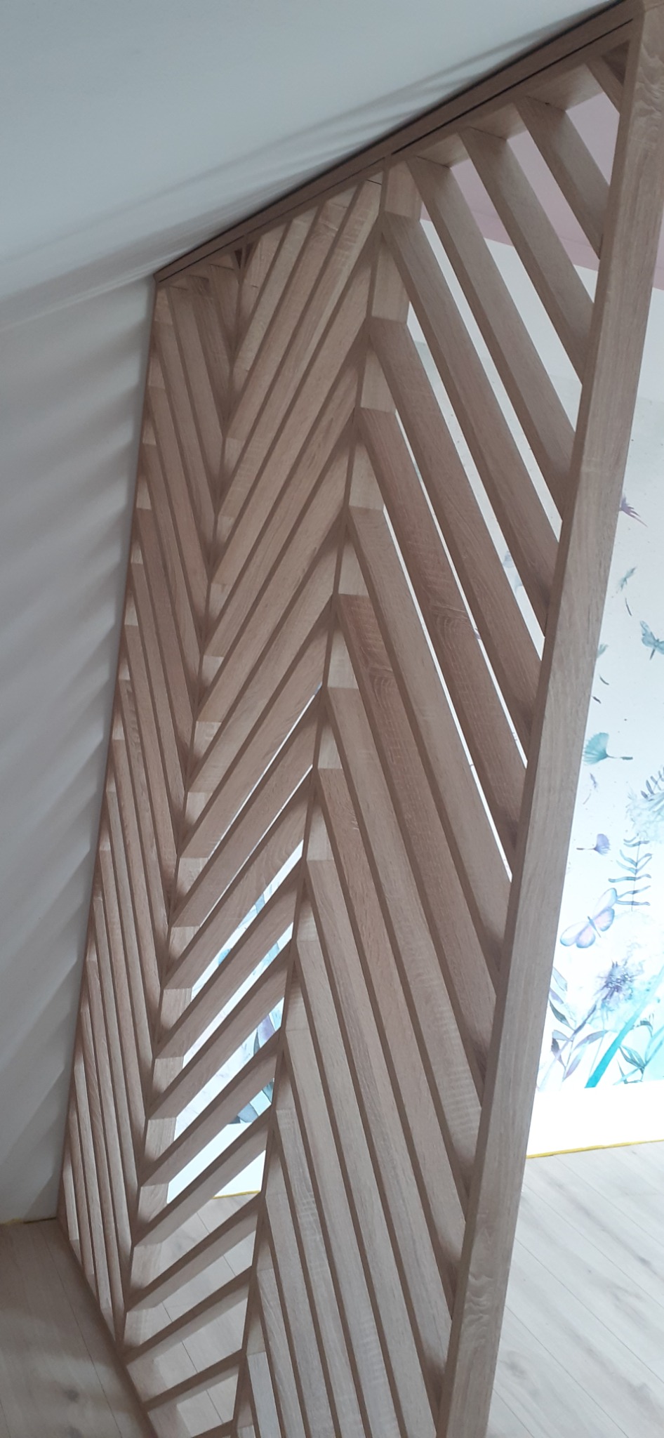 Drewniana ażurowa ścianka działowa z jasnego drewna, ułożona we wzór jodełki, oddzielająca przestrzeń z tapetą w kwiaty i motyle, widoczna z boku, na tle białej ściany i jasnej podłogi.