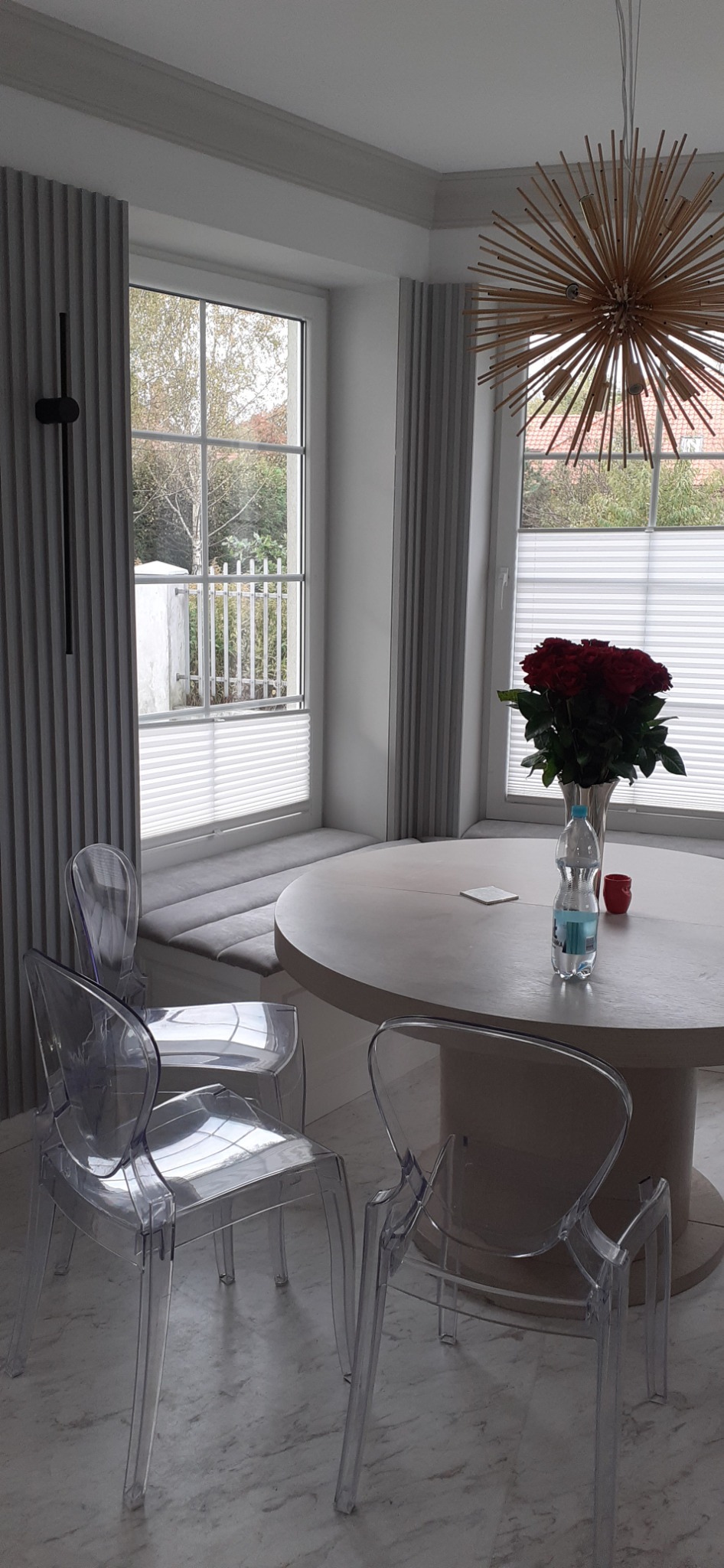 Jasne wnętrze jadalni z okrągłym stołem, bukietem czerwonych róż i przezroczystymi krzesłami; w tle okno z roletami plisowanymi i siedziskiem wnękowym.