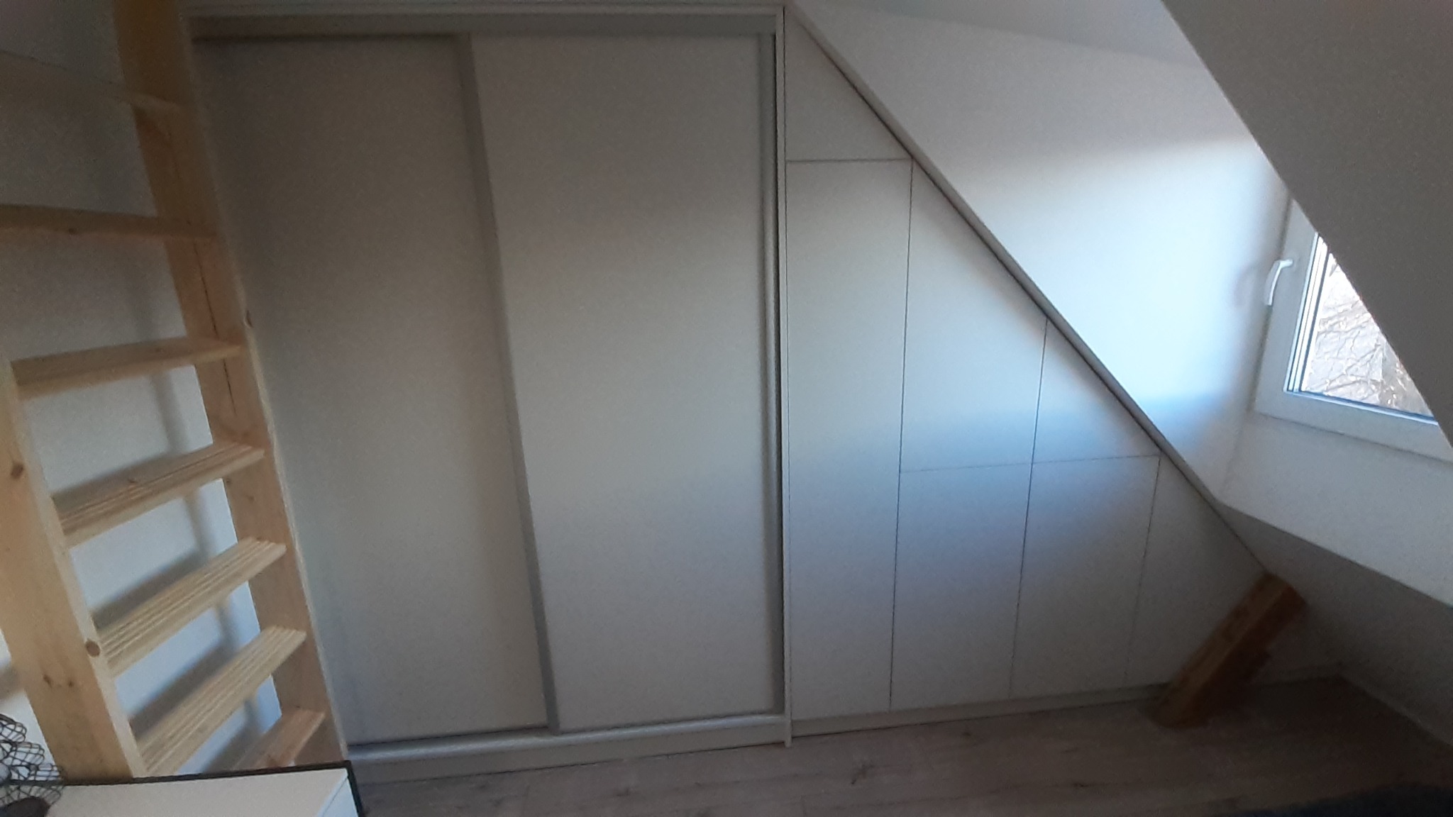 Biała szafa wnękowa z przesuwnymi drzwiami i drewnianą drabinką w pomieszczeniu na poddaszu w Olecku.