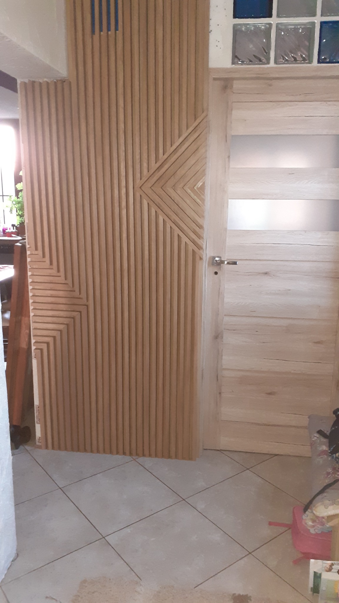 Drewniana ściana działowa z geometrycznym wzorem w pionowe lamele, sąsiadująca z jasnymi, drewnianymi drzwiami z przeszkleniem w górnej części; widoczne fragmenty otoczenia.