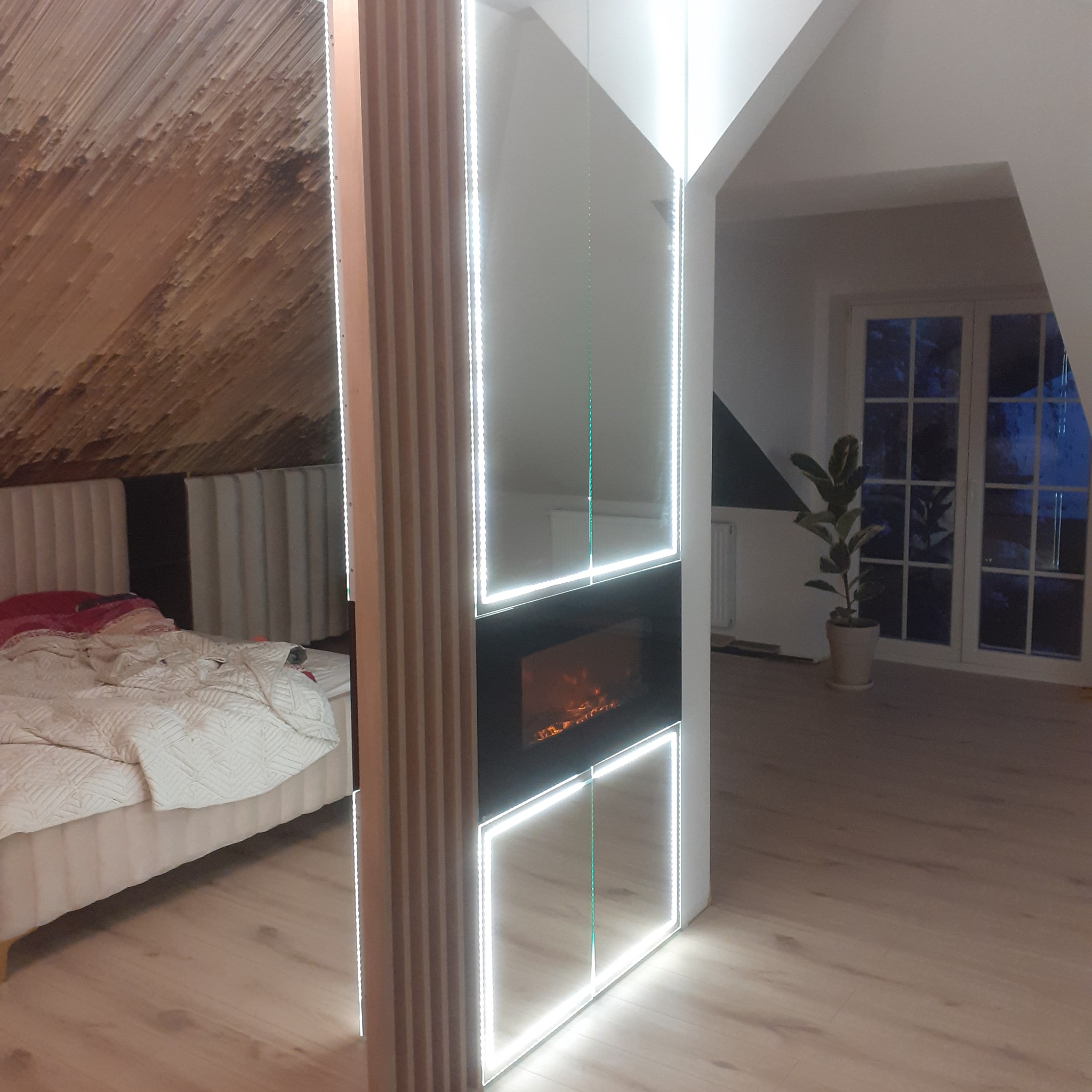 Nowoczesna sypialnia z łóżkiem i biokominkiem w zabudowie, oświetlonym listwami LED, na poddaszu z drewnianą boazerią na skosach.