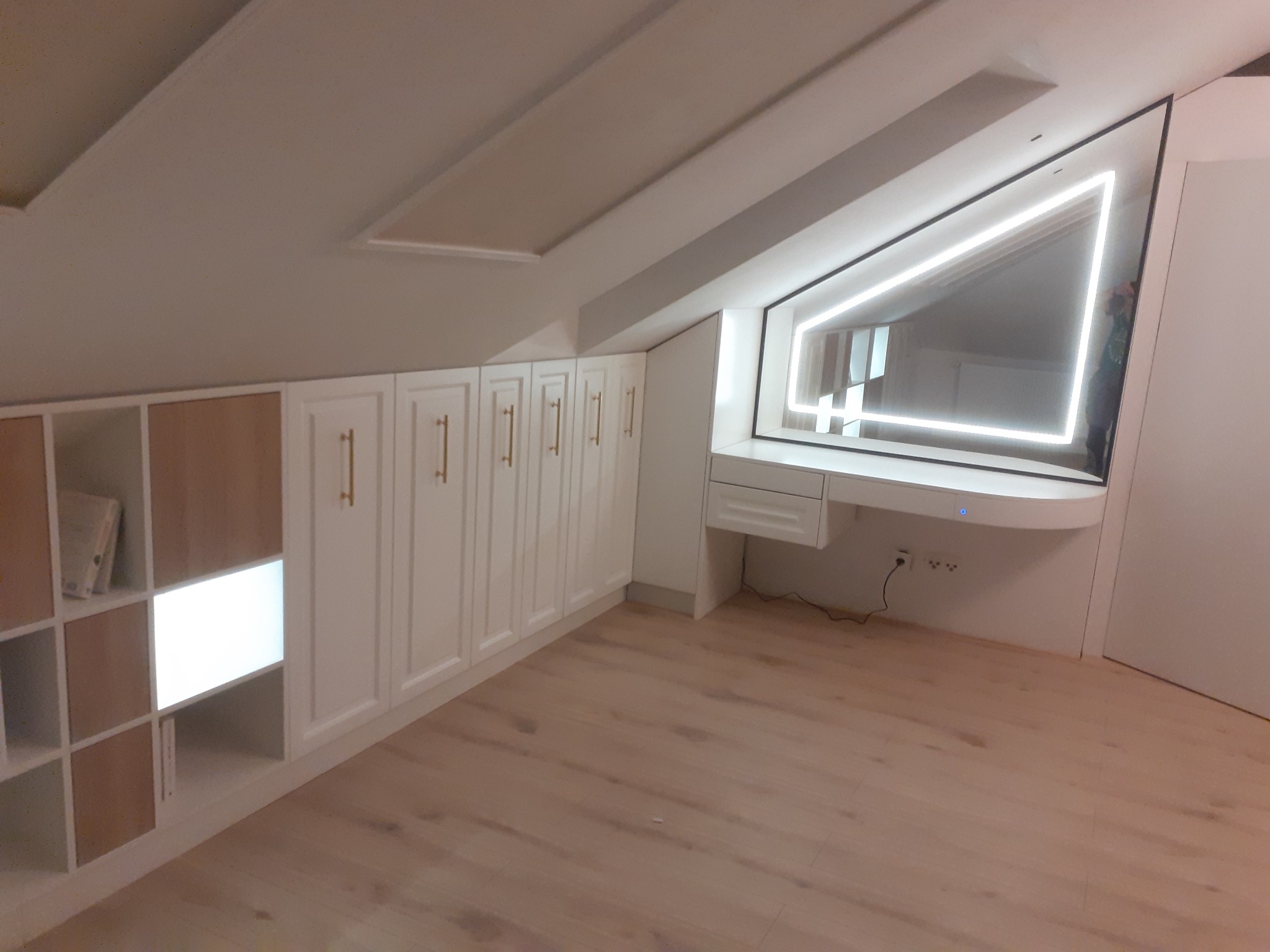 Białe szafy wnękowe z frezowanymi frontami i złotymi uchwytami w pomieszczeniu na poddaszu, połączone z toaletką z podświetlanym lustrem w kształcie trapezu.