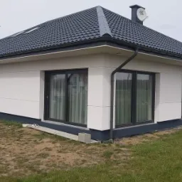 Nowoczesny, parterowy dom z szarą dachówką i białymi ścianami, widok z zewnątrz na elewację boczną z dużymi oknami i fragmentem ogrodu.