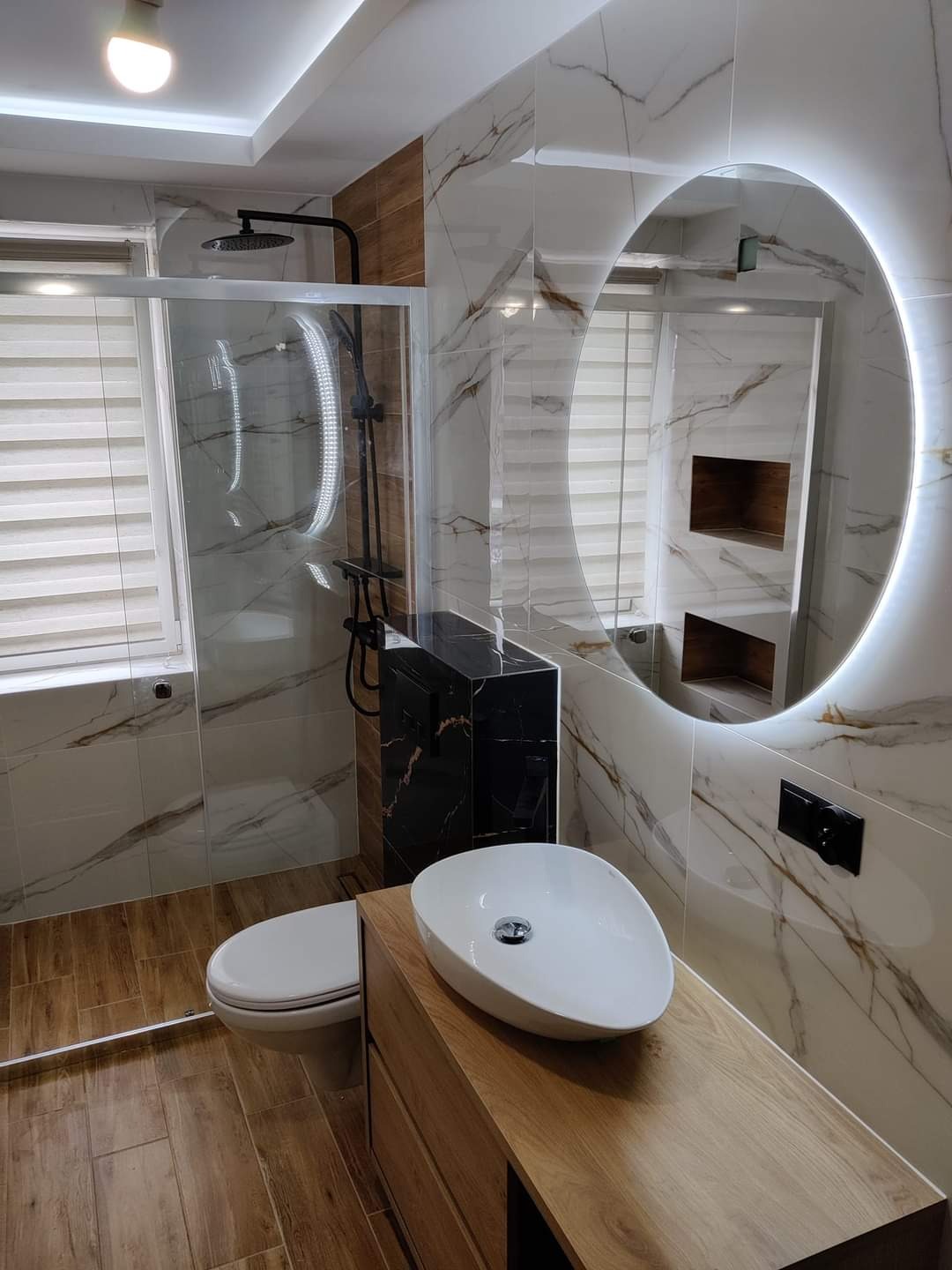 Nowoczesna łazienka z kabiną prysznicową typu walk-in, umywalką nablatową na drewnianej szafce, podświetlanym lustrem i marmurowymi płytkami z subtelnym żyłowaniem.