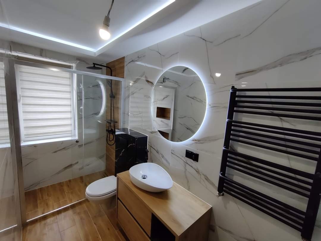 Nowoczesna łazienka z drewnianymi panelami podłogowymi, marmurowymi płytkami ściennymi, okrągłym lustrem z podświetleniem LED, czarnym grzejnikiem drabinkowym i kabiną prysznicową ze szklanymi...