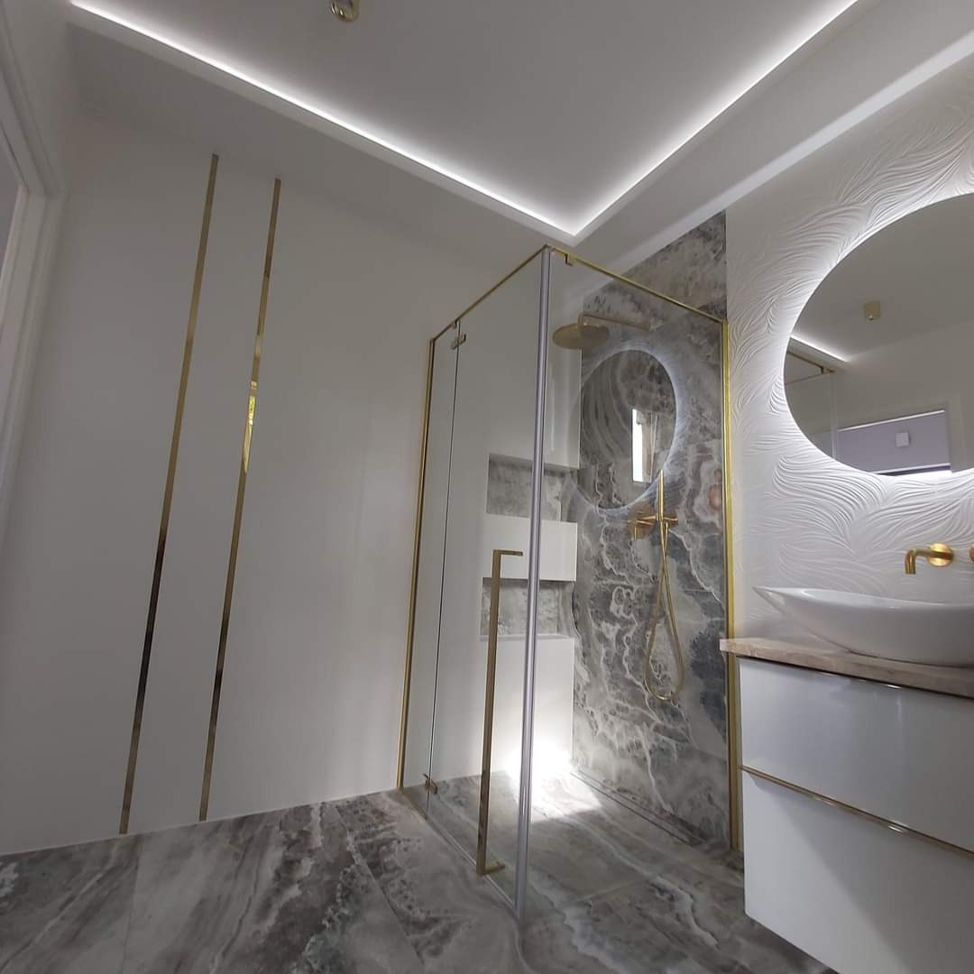 Nowoczesna łazienka z kabiną prysznicową ze złotymi detalami, okrągłym lustrem z podświetleniem LED, marmurową ścianą i szarą marmurową posadzką.