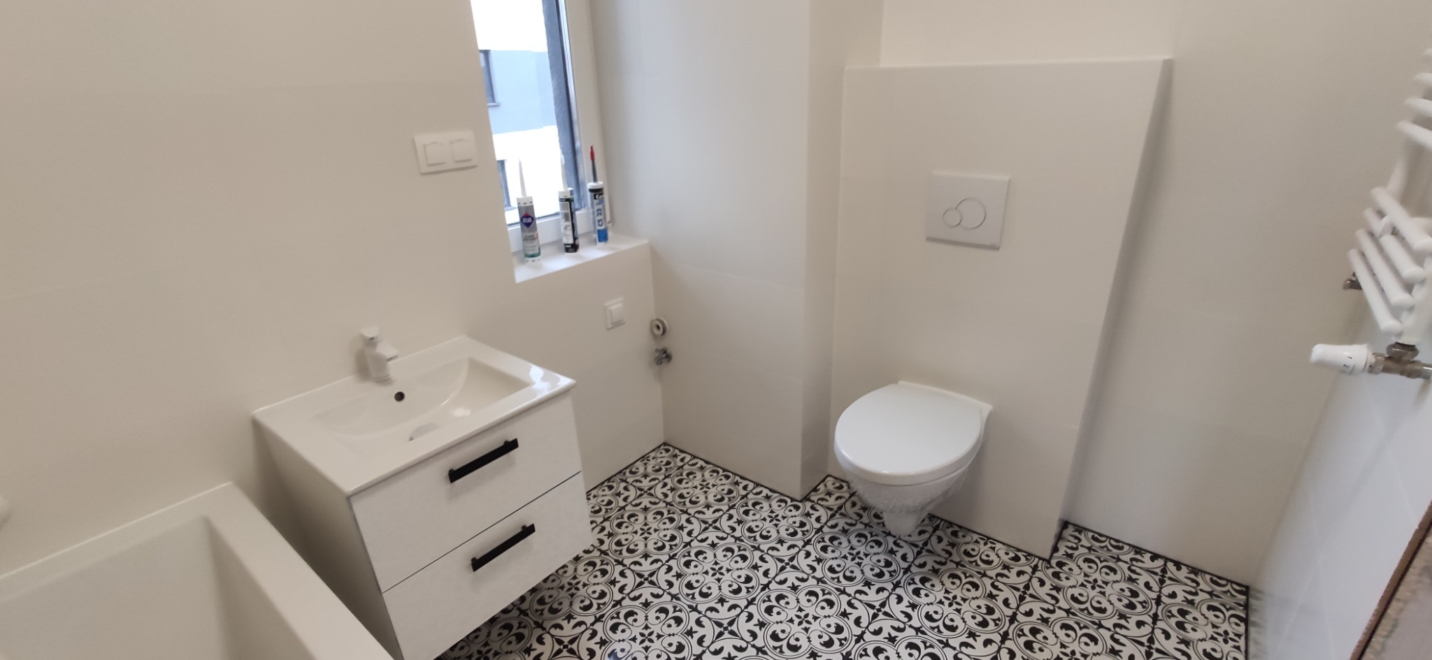 Biała łazienka z wzorzystą czarno-białą podłogą, umywalką z szafką, toaletą podtynkową i grzejnikiem drabinkowym, widoczne tuby z silikonem na parapecie.