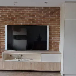 Nowoczesna zabudowa ściany z cegły: szafka RTV z drewnopodobnymi frontami, białe szafki wiszące z drapakiem dla kota oraz wysoka, jasna szafa wnękowa zlicowana ze ścianą.