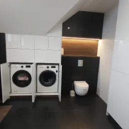 Nowoczesna łazienka z pralkami ukrytymi w białych szafkach na wymiar, czarne płytki podłogowe i ściana, toaleta w tle, oświetlenie LED pod szafką wiszącą.