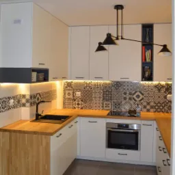 Nowoczesna kuchnia w kształcie litery U z białymi szafkami, drewnianym blatem, czarną armaturą i wzorzystą mozaiką na ścianie. Nad blatem zamontowane oświetlenie LED, a nad nim designerska, czarna...