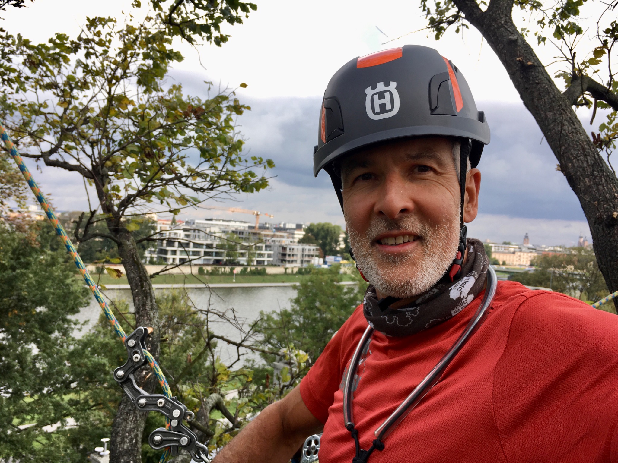 Arborysta uśmiecha się, w hełmie Husqvarna, zabezpieczony linami podczas pracy na drzewie z widokiem na miasto i rzekę w tle.