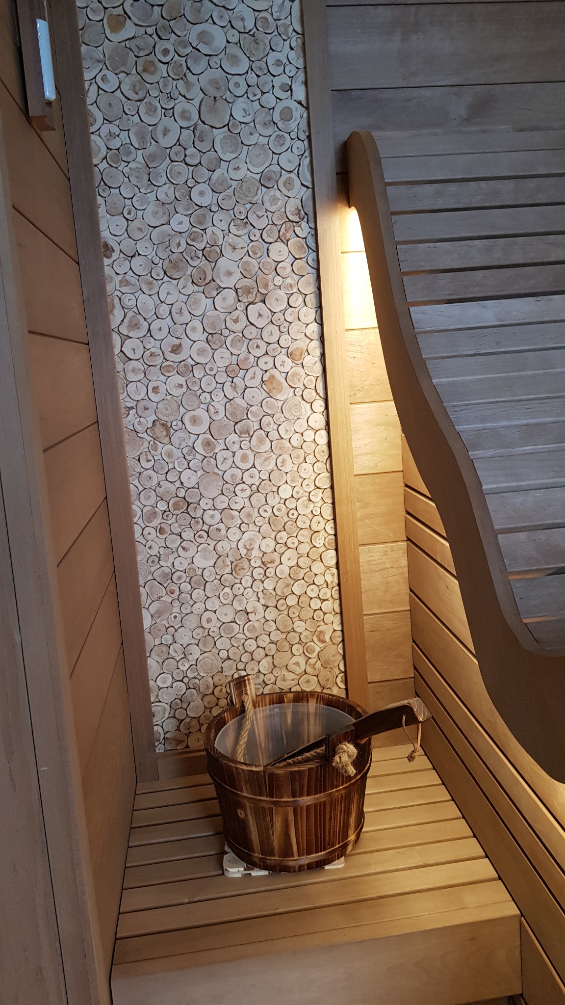 Wnętrze drewnianej kabiny z ławkami, dekoracyjną ścianą z plasterków drewna i drewnianym wiadrem z chochlą na podłodze.