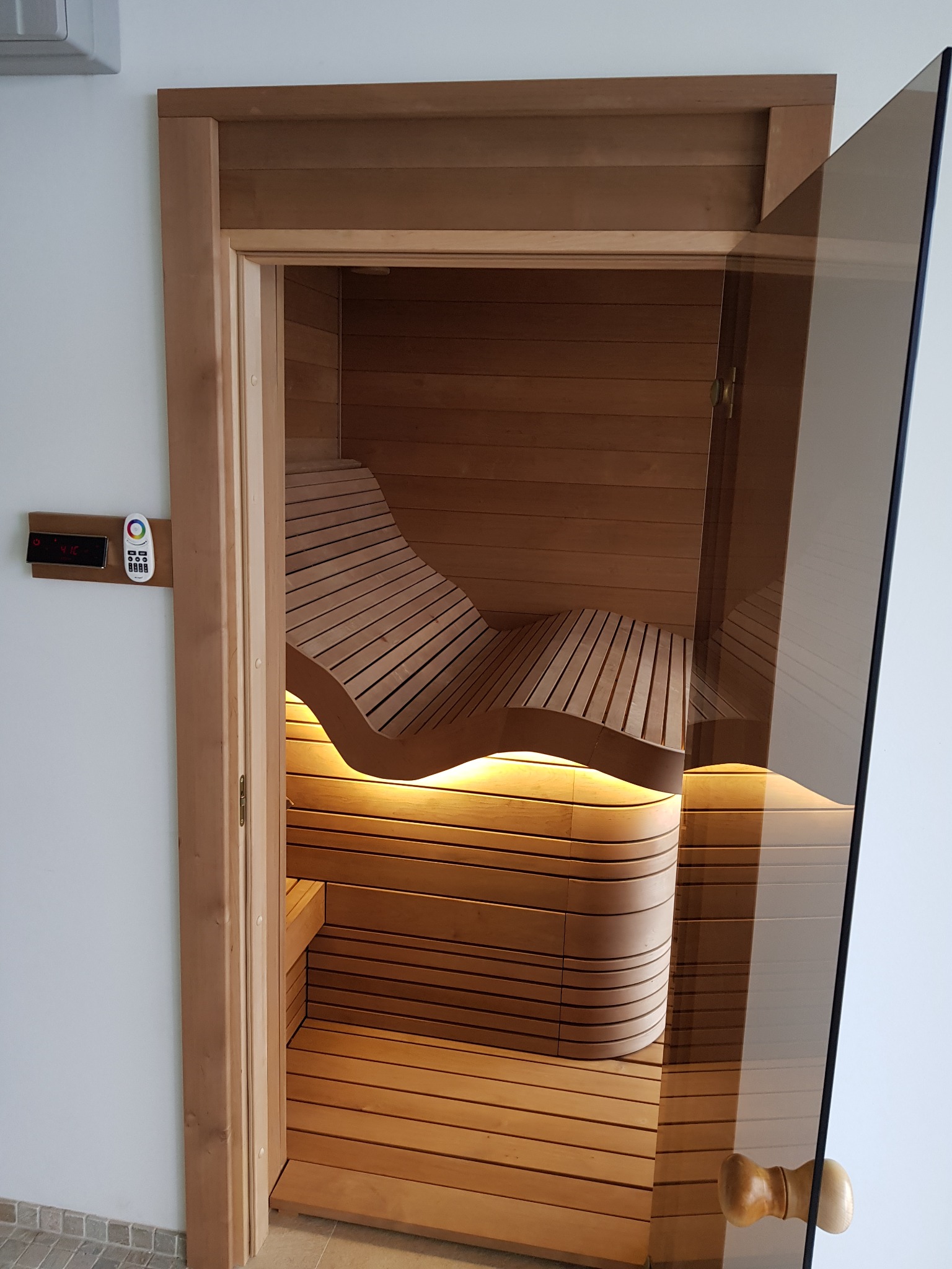 Nowoczesna kabina z drewna z podświetlanymi ławkami o falistym kształcie, widoczny panel sterowania i szklane drzwi.