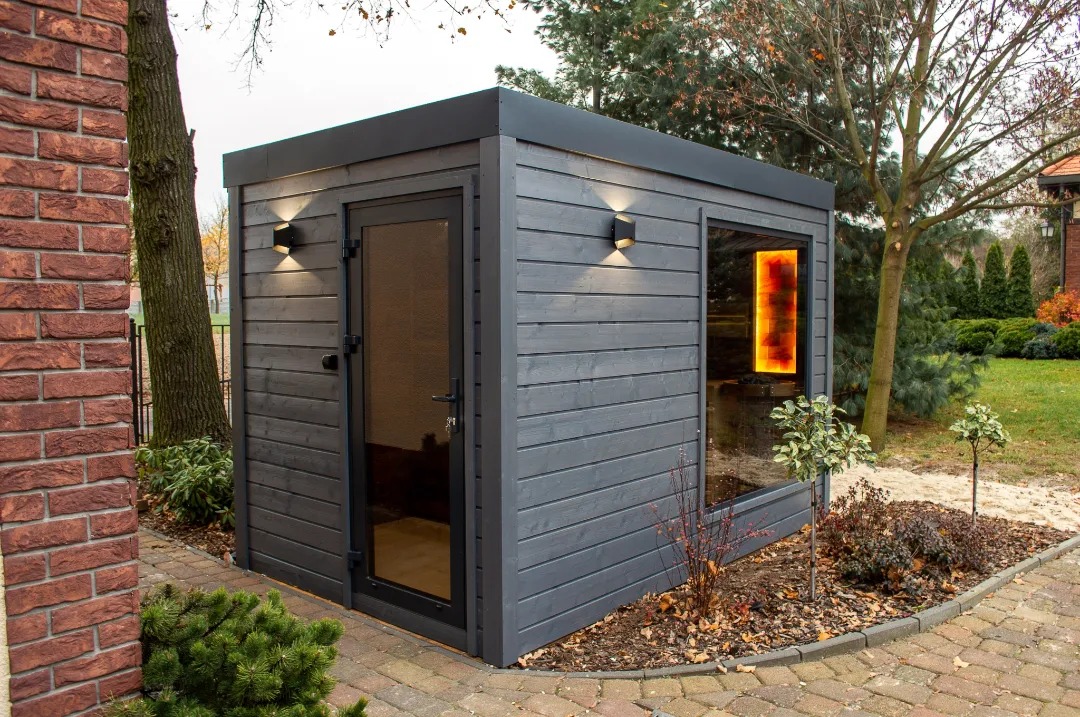 Nowoczesna, drewniana kabina z przeszklonym oknem, oświetlona dwoma kinkietami, umieszczona w ogrodzie obok ceglanego budynku.