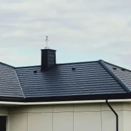 Czarny dach z kominem i rynną w Radomiu, widok z dołu na tle pochmurnego nieba.