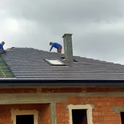 Montaż ciemnoszarej dachówki ceramicznej na nowym dachu z oknem dachowym i kominem z bloczków betonowych, widoczni dekarze w trakcie pracy.