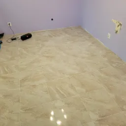 Ułożone beżowe płytki podłogowe w pokoju z fioletowymi ścianami, częściowo zakryte folią ochronną, widoczny głośnik przenośny i wystające elementy instalacji.