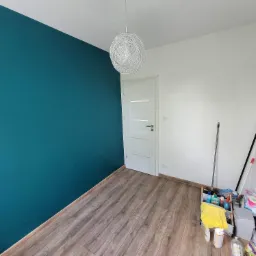 Świeżo pomalowany pokój z turkusową ścianą, drewnianą podłogą, nowoczesną lampą wiszącą i białymi drzwiami. W rogu widoczny wózek ze środkami czystości.
