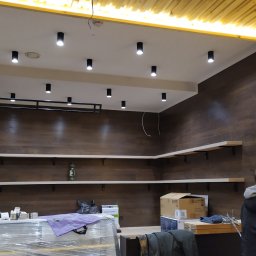 liberty live - Wnętrze biura w trakcie wykańczania: drewnopodobne panele ścienne, białe półki, oświetlenie punktowe sufitowe i lampa wisząca, widoczne materiały budowlane i osoby.