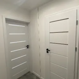 Dwa białe, nowoczesne drzwi wewnętrzne z czarnymi klamkami i poziomymi wstawkami, ustawione w korytarzu o szarym podłożu i białych ścianach.