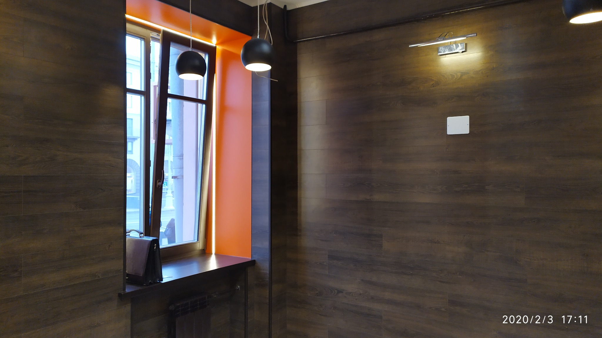 Wnętrze pokoju z drewnianą okładziną ścienną, oknem z pomarańczową wnęką i wiszącymi lampami kulistymi, uzupełnione o czarną torbę na parapecie.