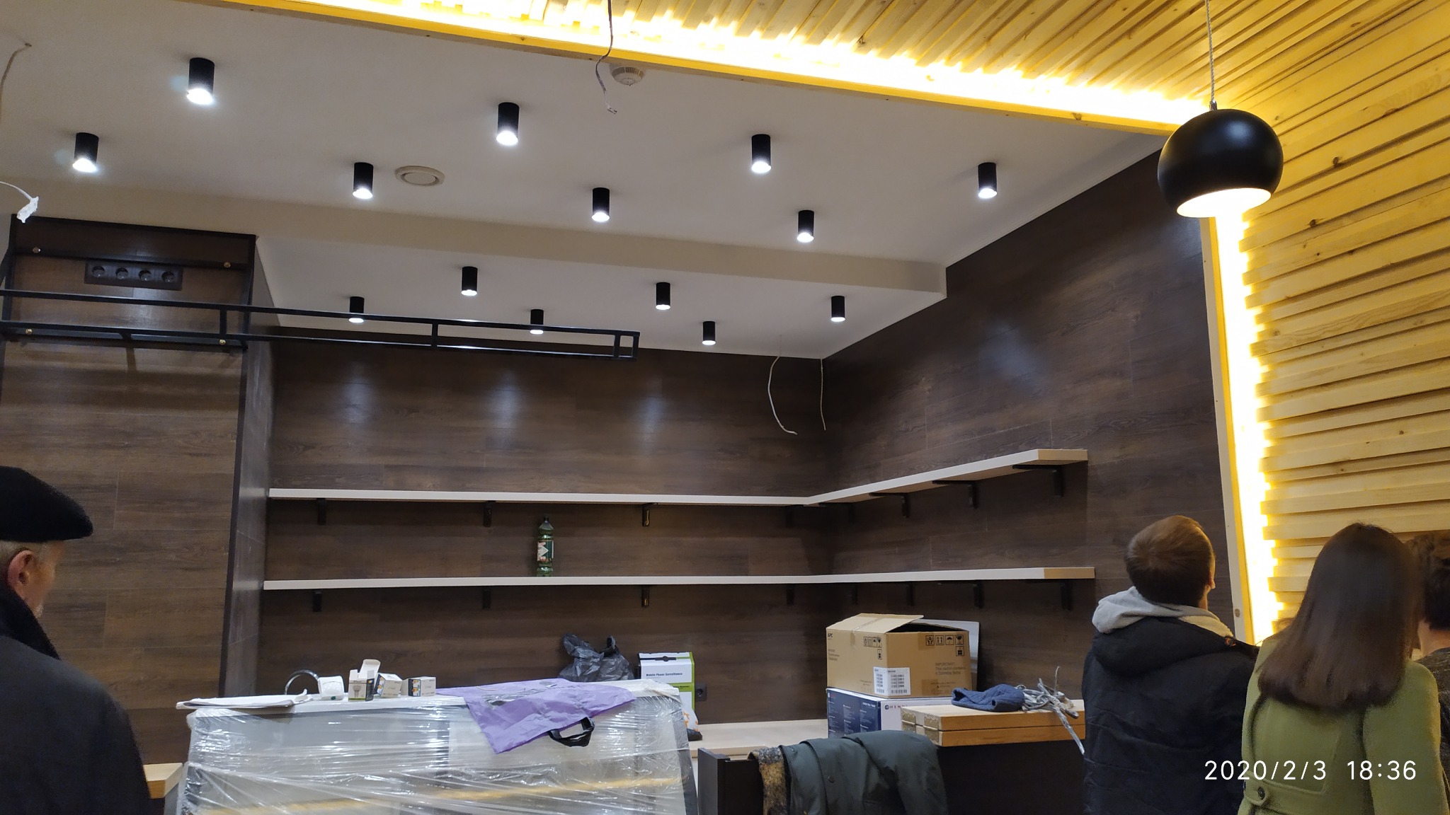 Wnętrze biura w trakcie wykańczania: drewnopodobne panele ścienne, białe półki, oświetlenie punktowe sufitowe i lampa wisząca, widoczne materiały budowlane i osoby.