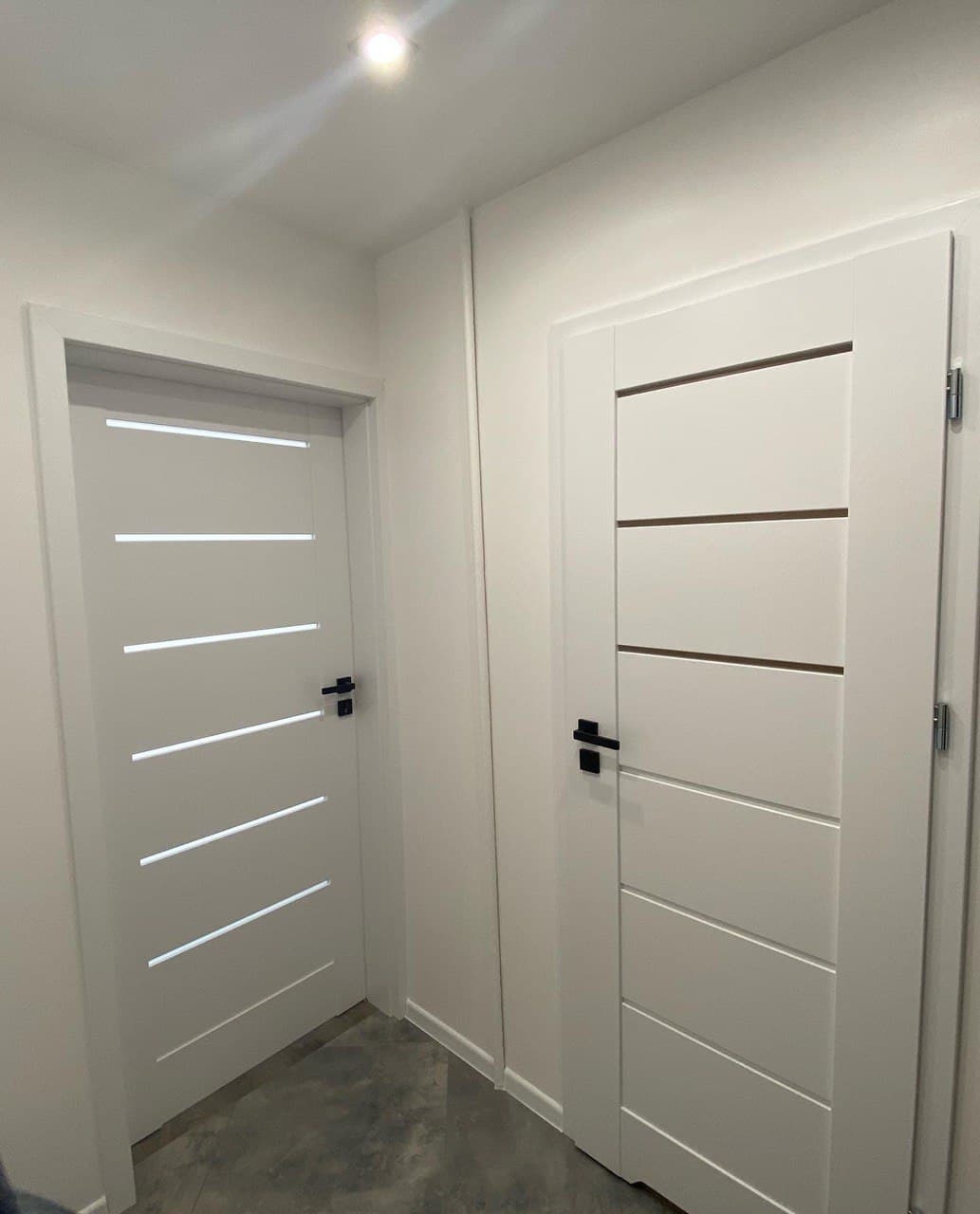 Dwa białe, nowoczesne drzwi wewnętrzne z czarnymi klamkami i poziomymi wstawkami, ustawione w korytarzu o szarym podłożu i białych ścianach.