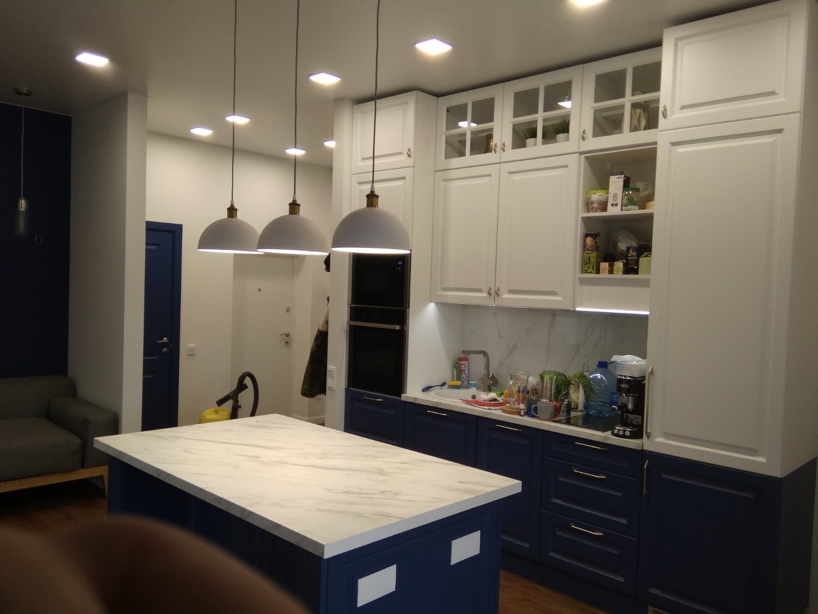 Nowoczesna kuchnia z wyspą o marmurowym blacie, granatowymi szafkami dolnymi i białymi górnymi, oświetlona wiszącymi lampami w kształcie kopuł.