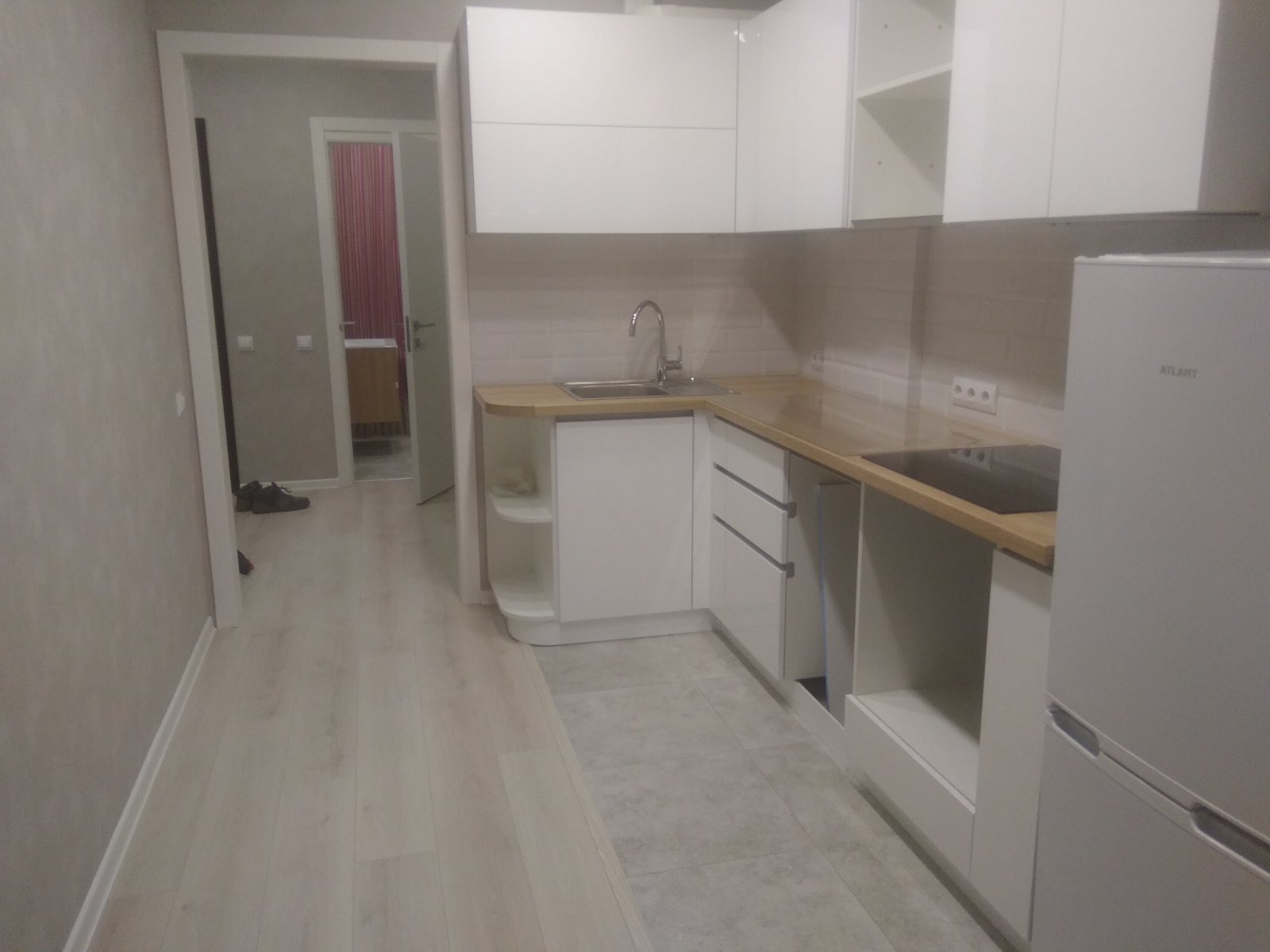 Nowoczesna, biała zabudowa kuchenna z drewnianym blatem, widoczny zlew, płyta indukcyjna i lodówka Atlant, z perspektywy wejścia do pomieszczenia.