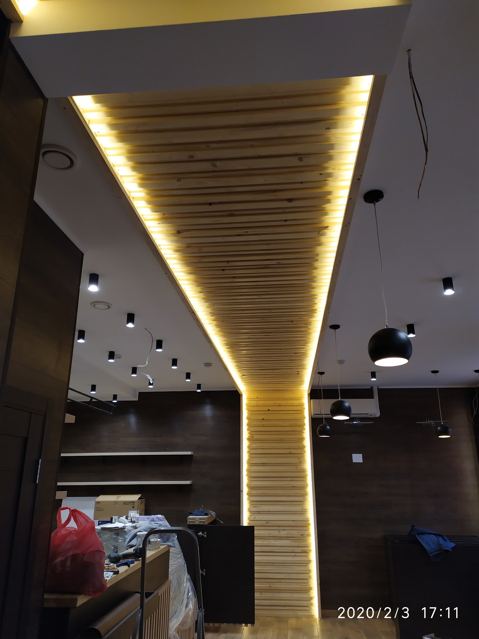 Podwieszany sufit z drewnianych listew z oświetleniem LED wzdłuż krawędzi, widoczne czarne lampy wiszące i punktowe oświetlenie sufitowe, fragment pomieszczenia w trakcie prac wykończeniowych.