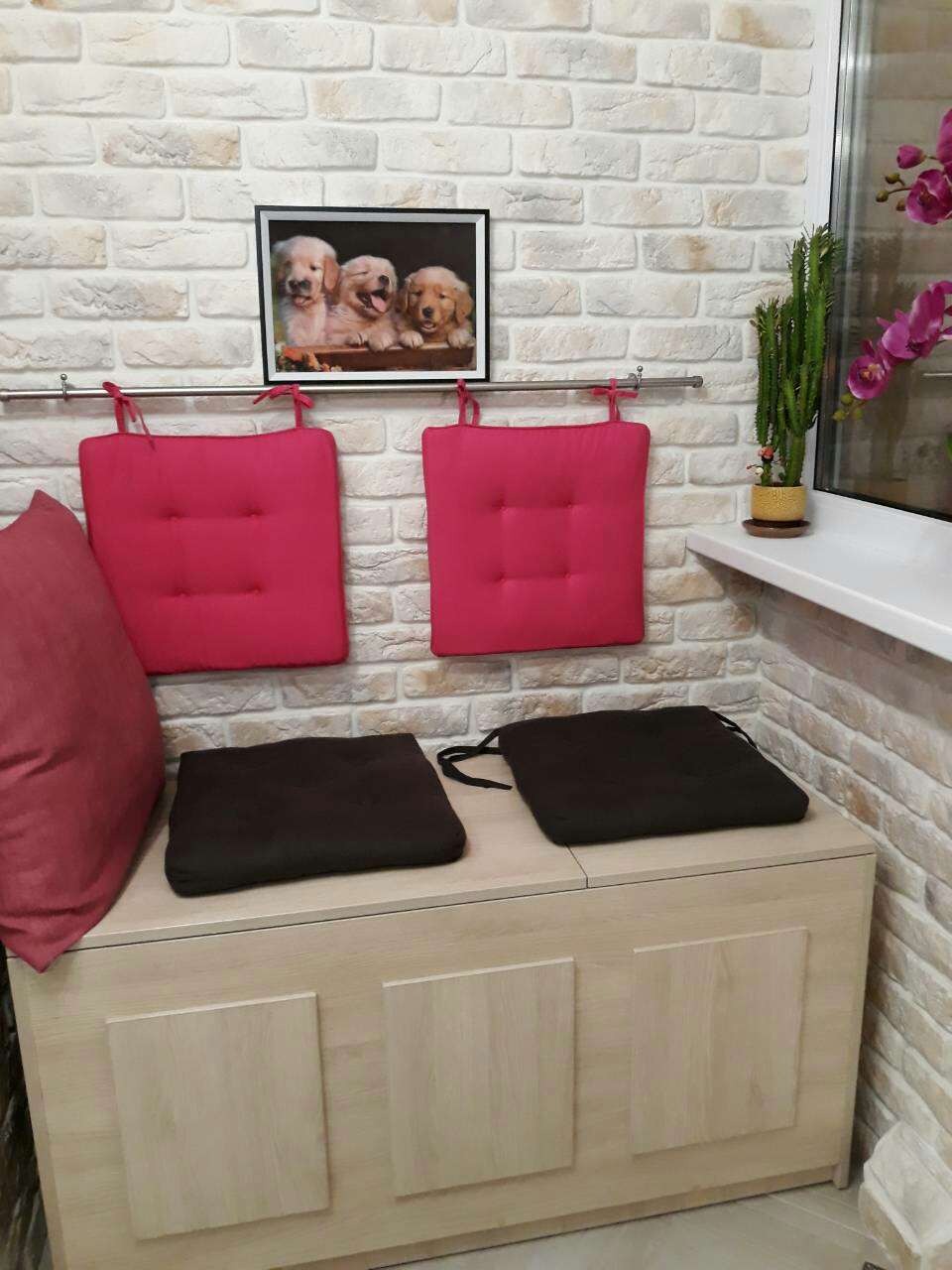 Jasny pokój z siedziskiem ze schowkiem, różowymi poduszkami wiszącymi na metalowym relingu i czarnymi poduszkami na siedzisku, na ścianie wisi obrazek z trzema szczeniakami rasy golden retriever.