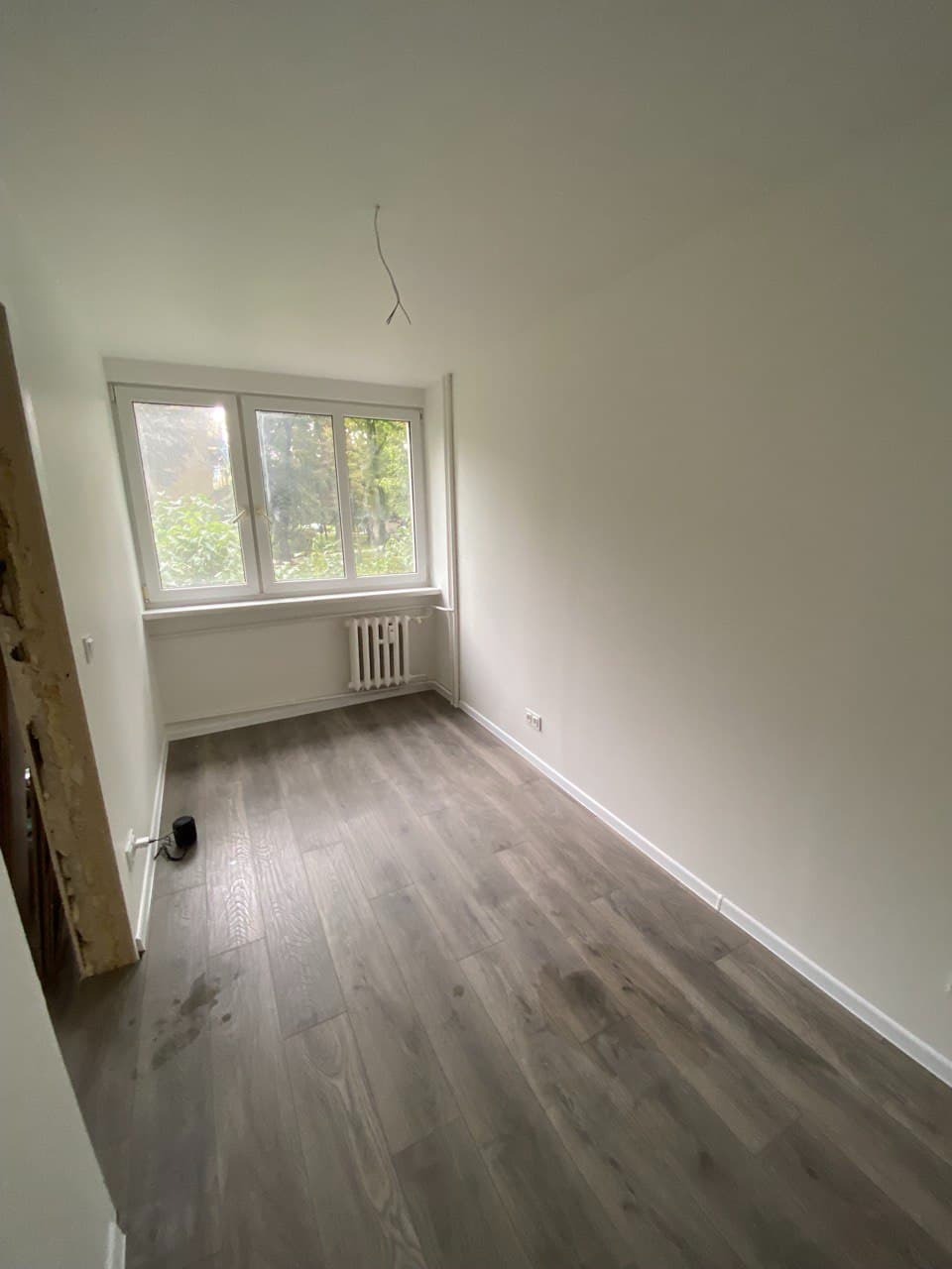 Pokój po odświeżeniu: szare panele podłogowe, świeżo pomalowane białe ściany i sufit, widoczny grzejnik pod oknem i wystający kabel ze ściany.