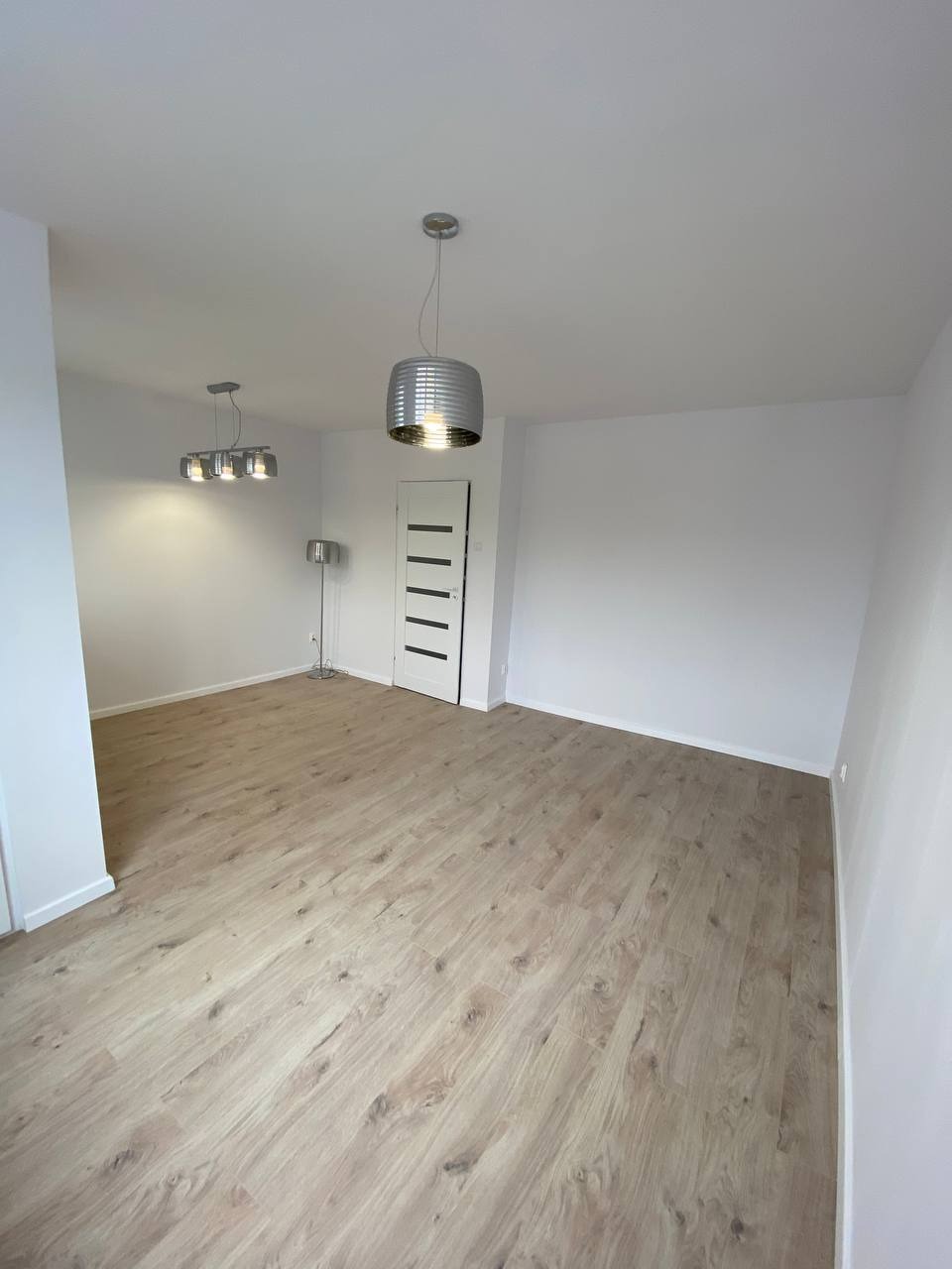 Wnętrze pokoju z panelami podłogowymi w odcieniach beżu i szarości, widoczne lampy sufitowe w stylu industrialnym i białe ściany z drzwiami.