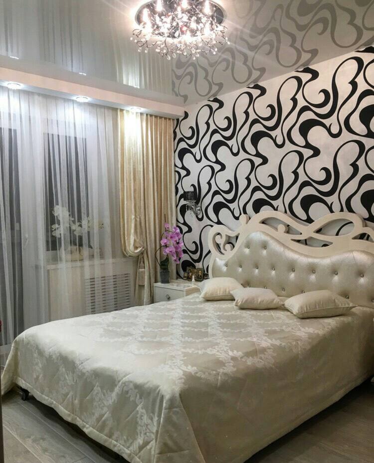 Elegancka sypialnia z błyszczącym sufitem napinanym, wzorzystą tapetą w czarno-białe fale i kremowym łóżkiem z pikowanym zagłówkiem, oświetlona ozdobnym żyrandolem.