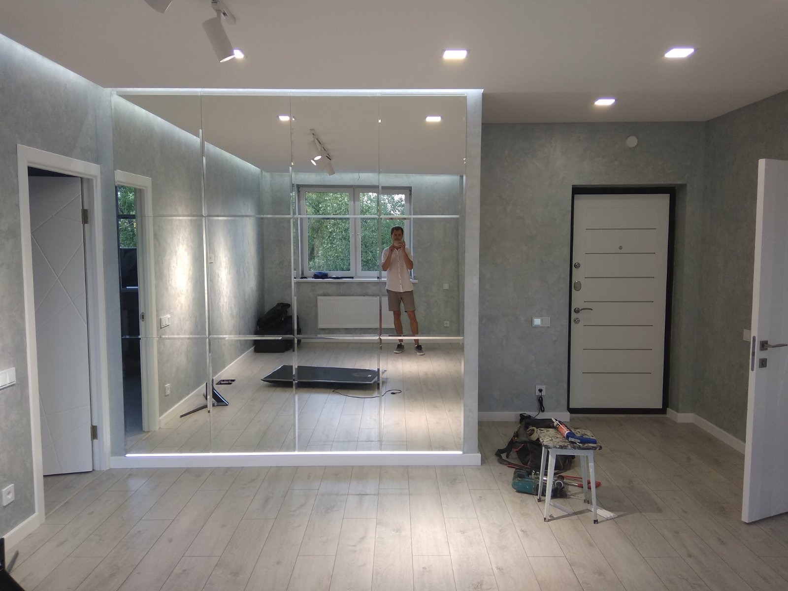 Duże lustro ścienne składające się z kwadratowych paneli, zamontowane w szarej ścianie, z widocznym odbiciem osoby robiącej zdjęcie telefonem i sprzętu do ćwiczeń w tle.