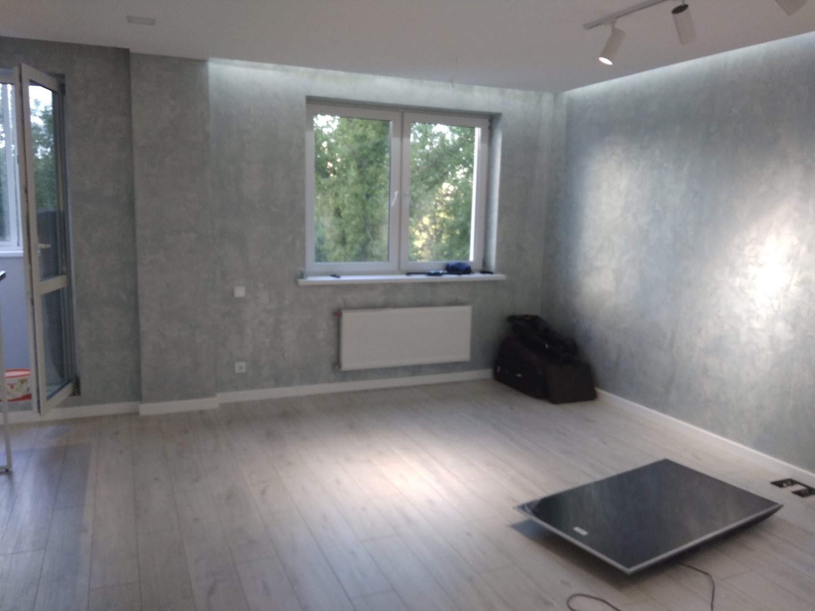 Pusty pokój z wykończonymi ścianami w odcieniach szarości, jasną podłogą, oknem z widokiem na zieleń i czarną płytą na podłodze, prawdopodobnie elementem wyposażenia lub narzędziem.