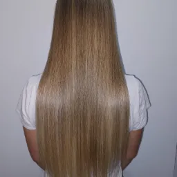 Długie, proste, blond włosy z ciemniejszym odrostem, widoczne z tyłu na tle białej ściany, po zabiegu prostowania.