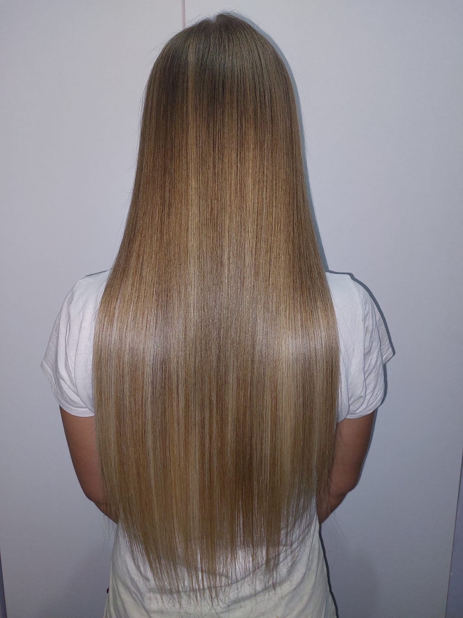 Długie, proste, blond włosy z ciemniejszym odrostem, widoczne z tyłu na tle białej ściany, po zabiegu prostowania.