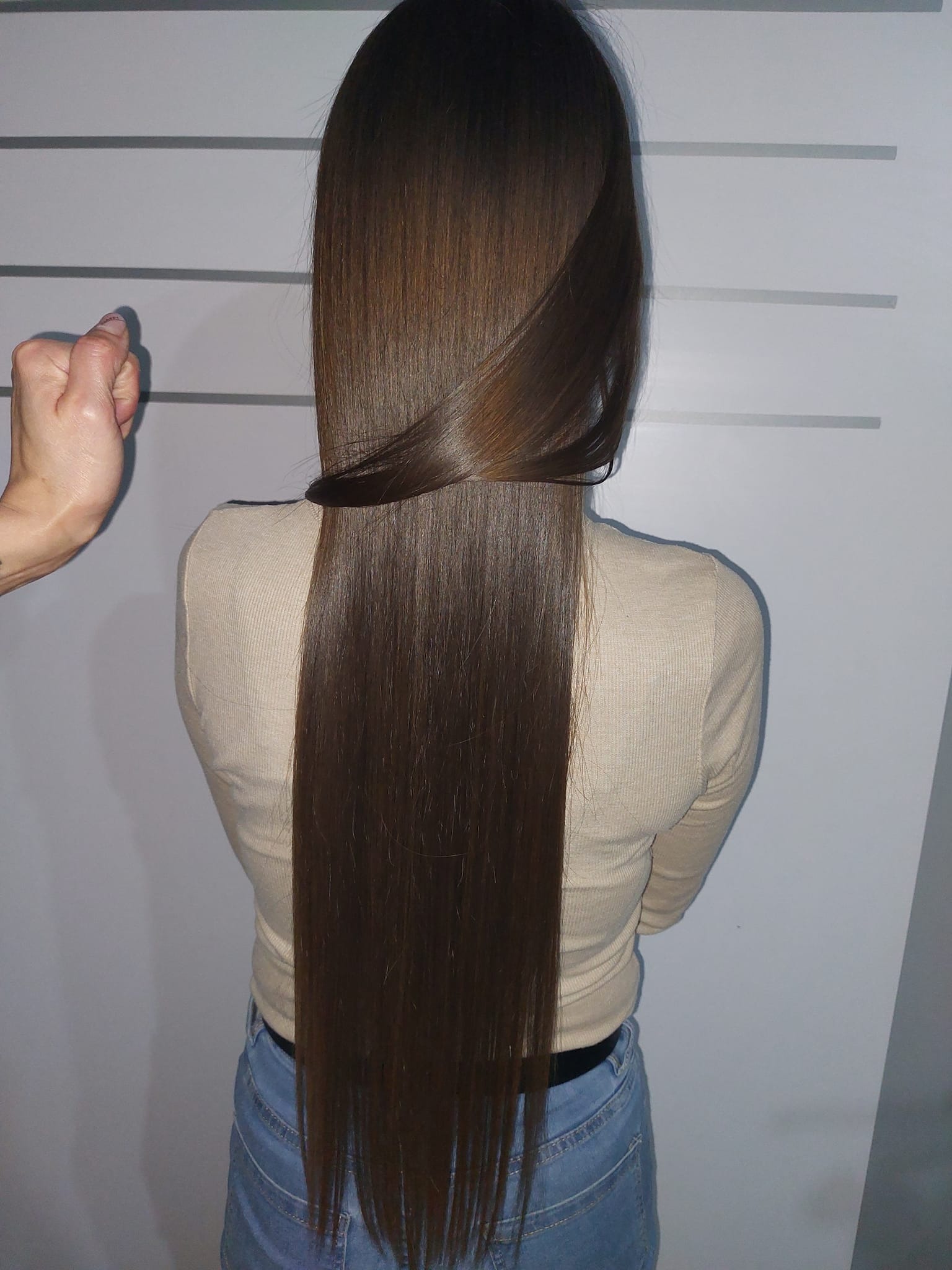 Widok z tyłu na bardzo długie, proste, brązowe włosy po zabiegu prostowania keratynowego. W tle jasnoszara ściana z poziomymi liniami. W lewym górnym rogu widoczna dłoń.