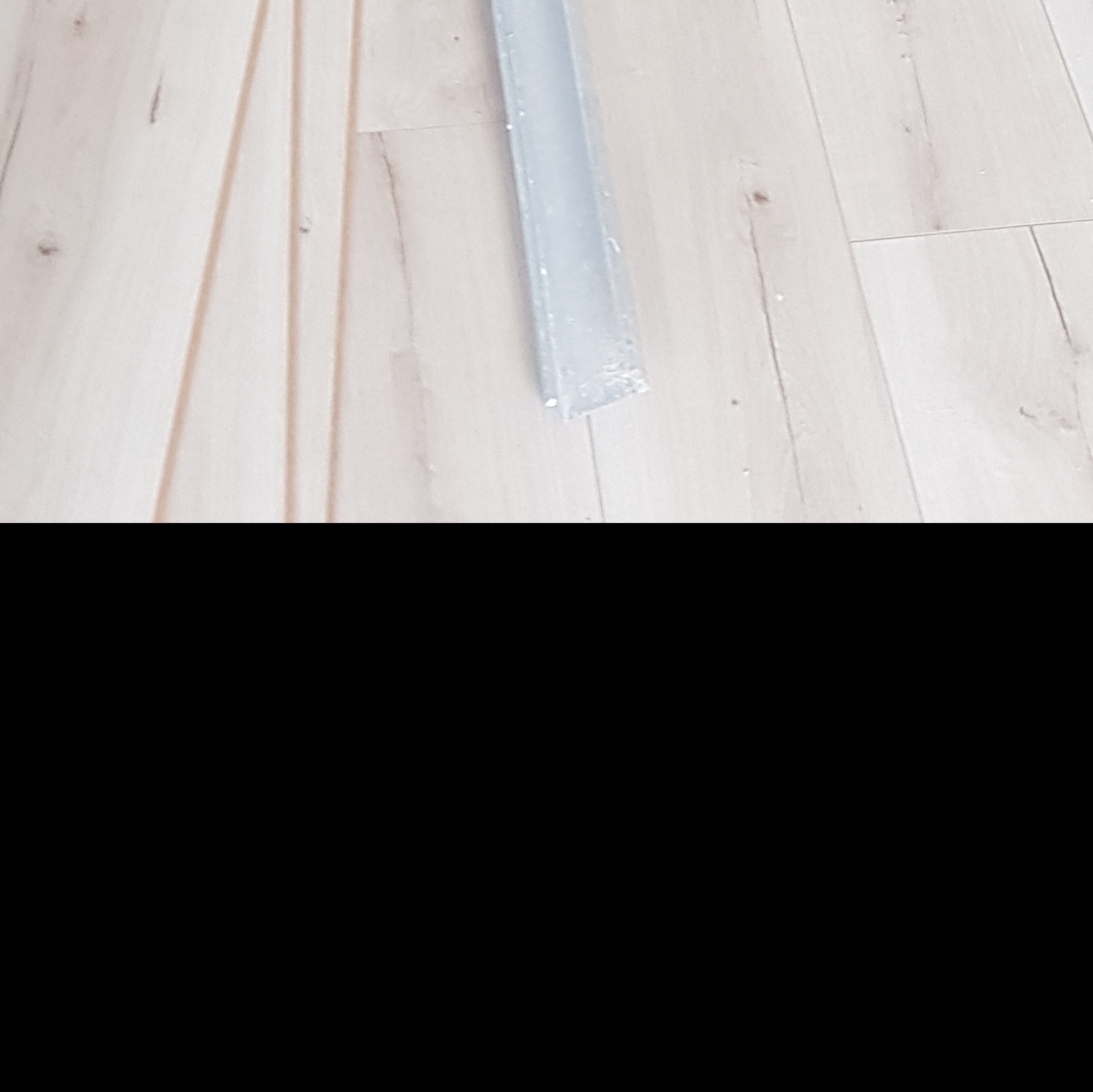 Fragment podłogi z jasnego drewna z widocznymi listwami przypodłogowymi oraz profilem aluminiowym w trakcie montażu.