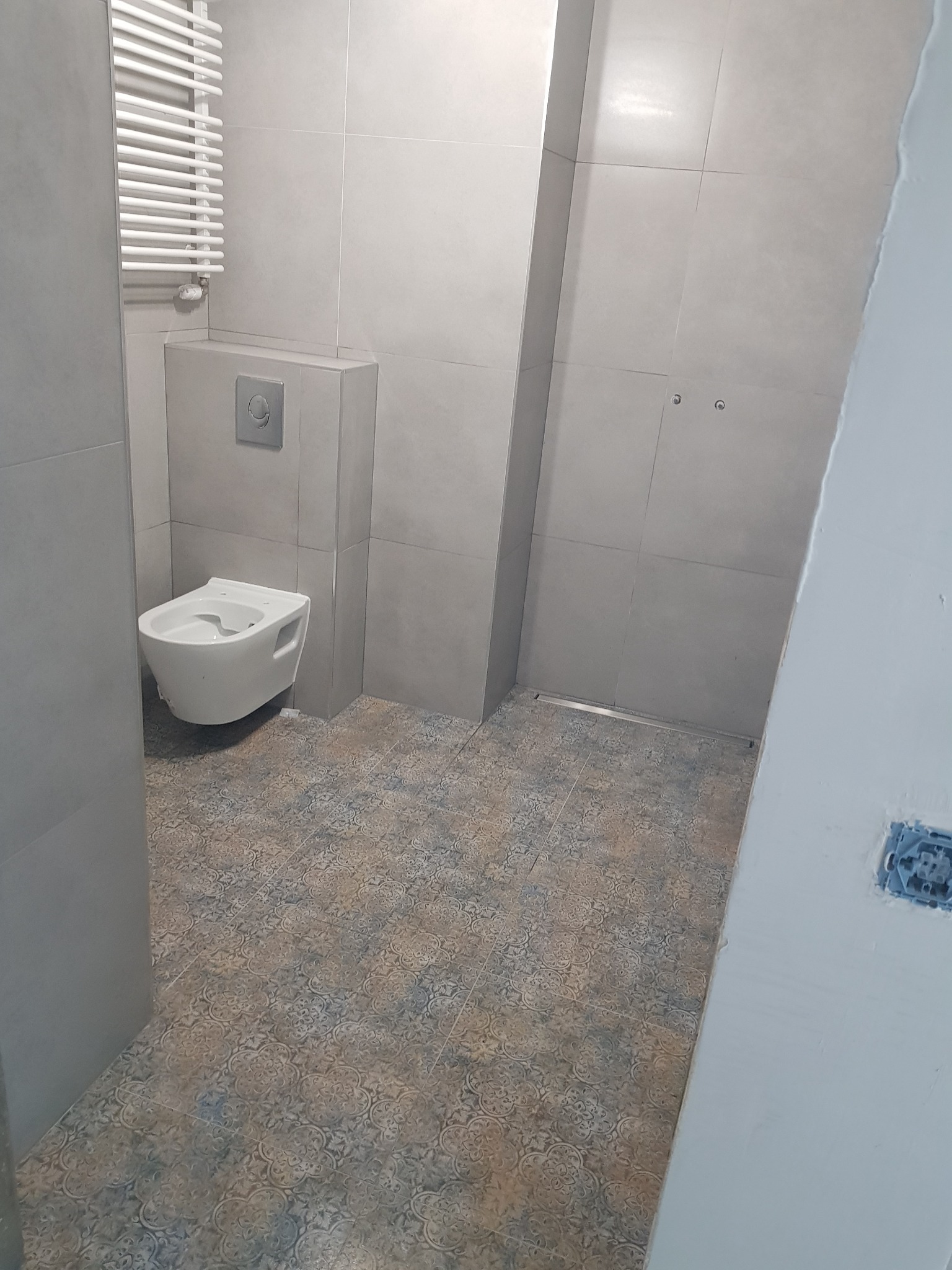 Nowoczesna łazienka z podwieszaną toaletą, grzejnikiem drabinkowym i płytkami podłogowymi w stylu vintage, widok z rogu pomieszczenia.