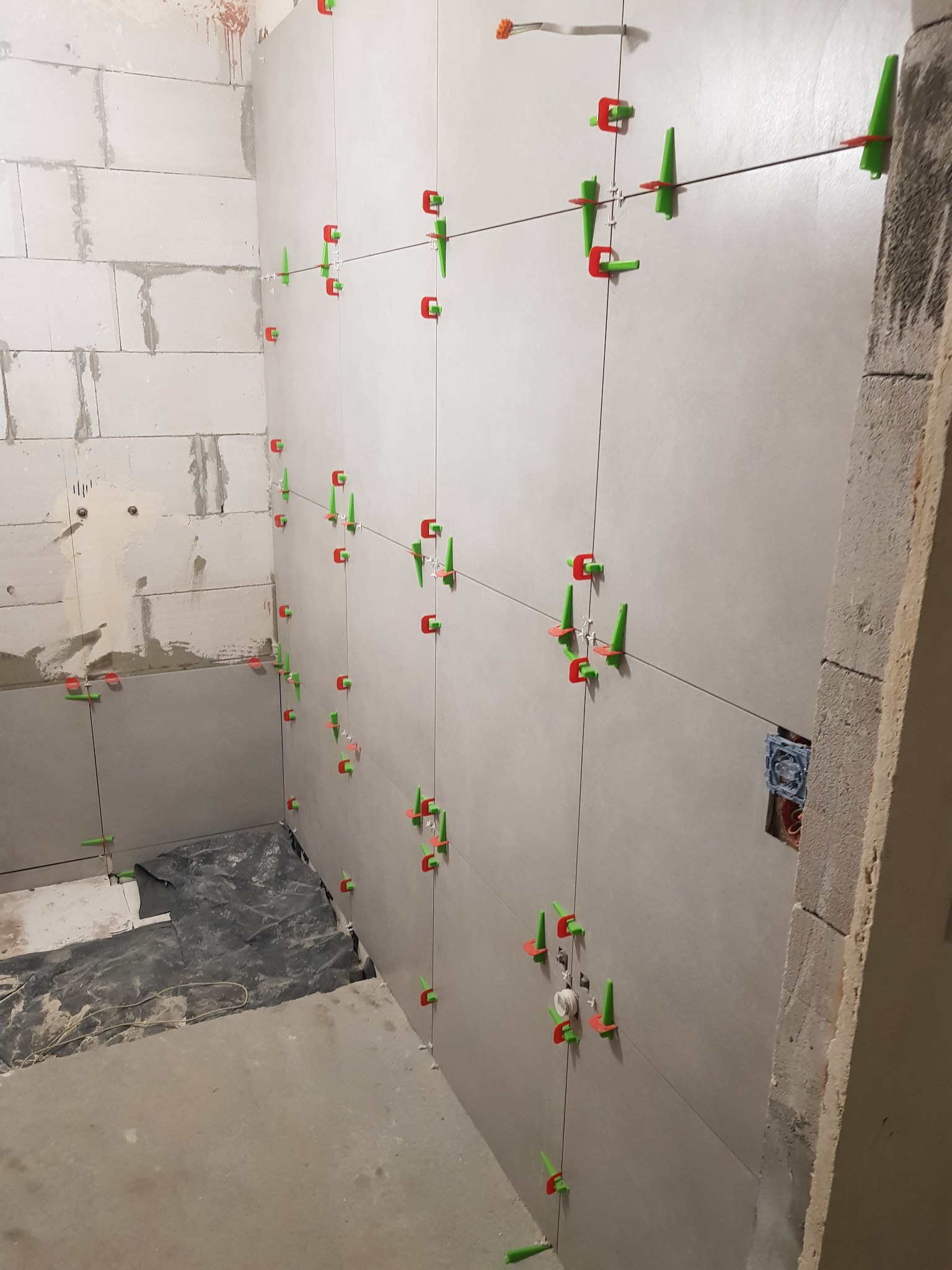 Świeżo ułożone, jasnoszare płytki na ścianie w trakcie kładzenia, z użyciem czerwono-zielonych klipsów poziomujących, widoczne surowe ściany z betonu komórkowego i przygotowane podłoże na podłodze.