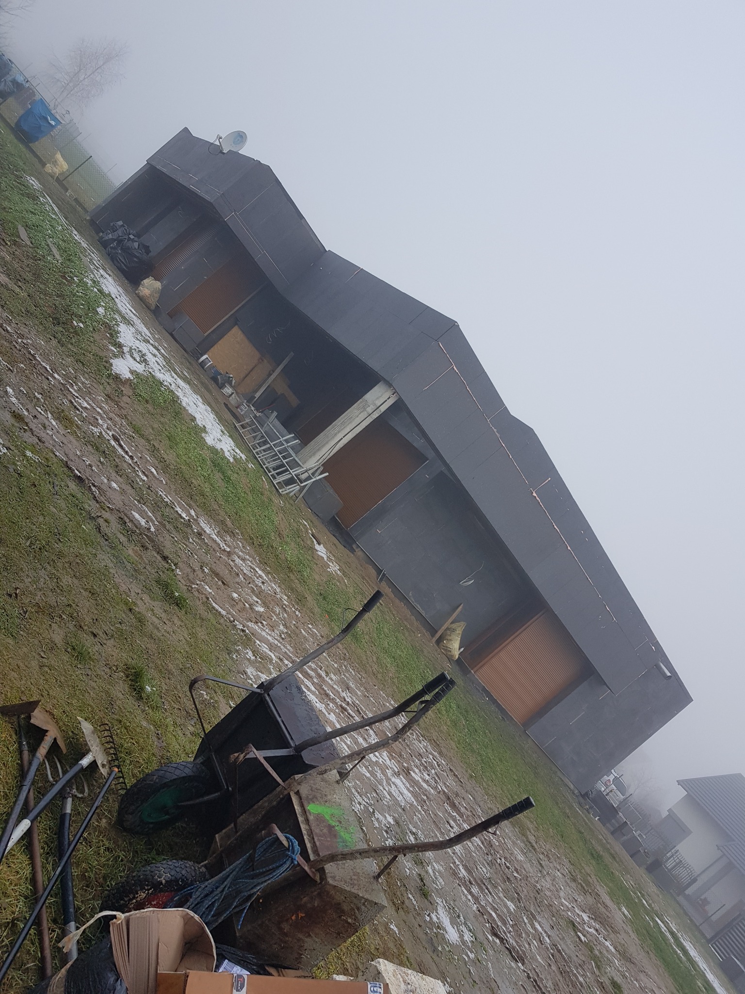 Budynek w trakcie ocieplania ścian zewnętrznych, widoczny w mglisty dzień z narzędziami budowlanymi na pierwszym planie.