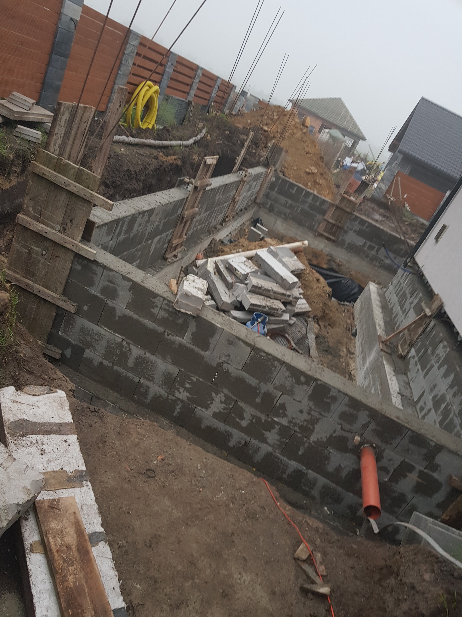 Fundamenty domu w trakcie budowy, widoczne bloczki betonowe, szalunki drewniane, rury kanalizacyjne oraz ziemia i ogrodzenie w tle.