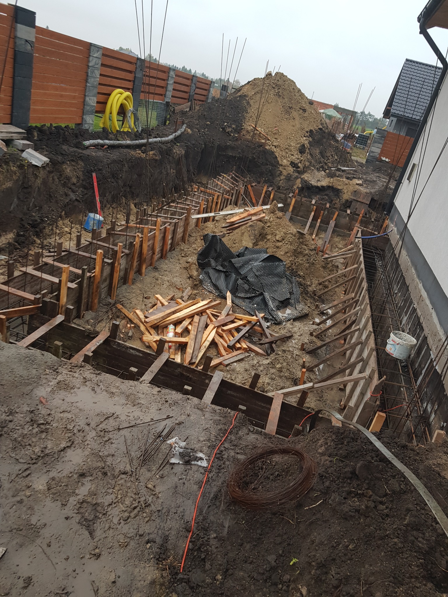 Wykop fundamentów z deskowaniem i zbrojeniem, widoczny stos drewna, zwój drutu wiązałkowego, oraz żółte rury w tle.