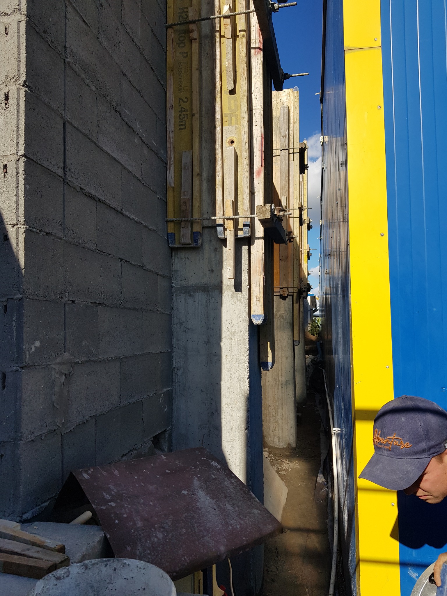 Szare bloczki fundamentowe z widoczną konstrukcją szalunkową i betonowymi słupami, częściowo zasłonięte blachą i fragmentem niebiesko-żółtej ściany kontenera budowlanego.