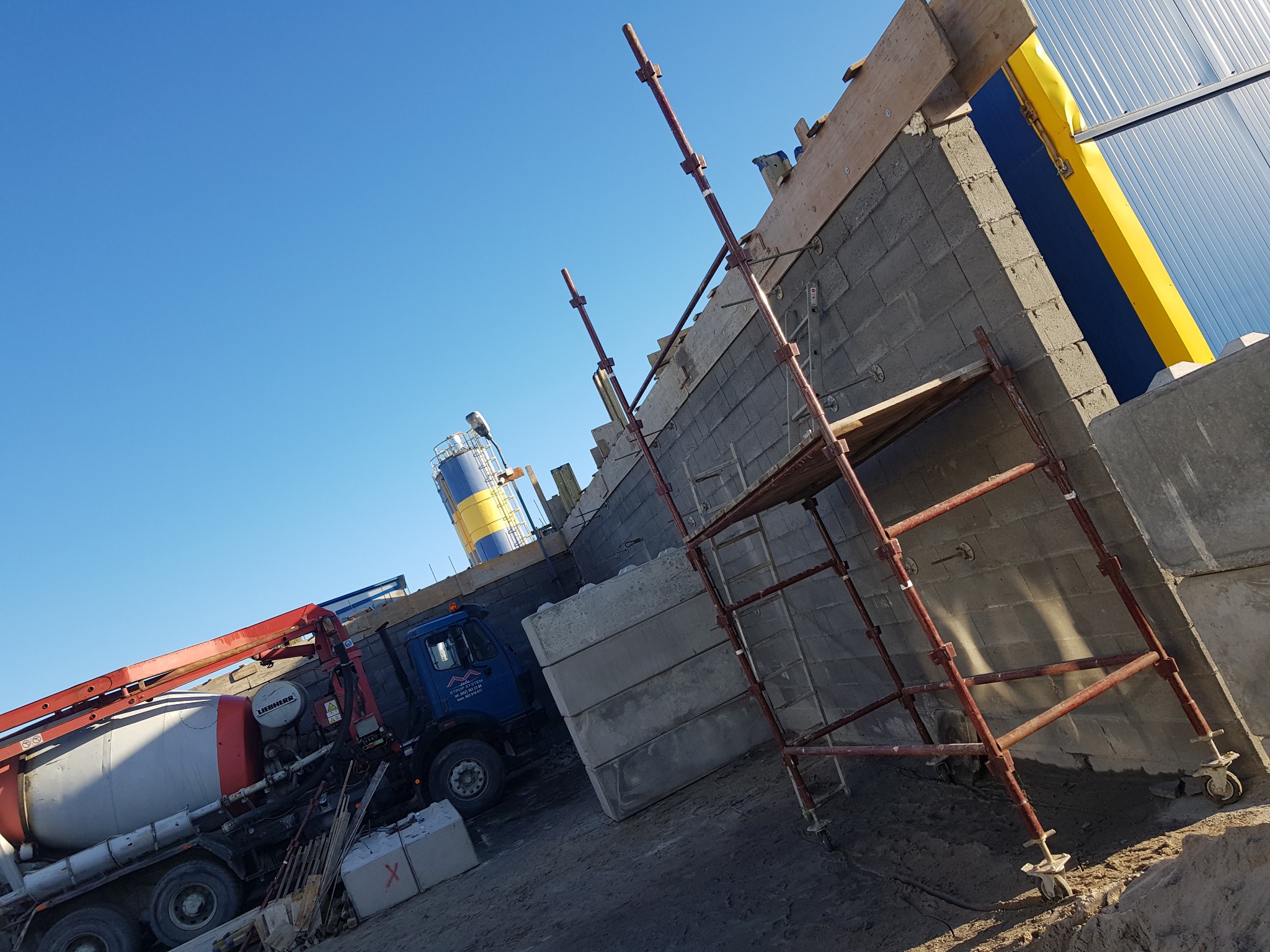 Budowa domu z bloczków betonowych, widoczny rusztowanie, betoniarka Liebherr i niebieski silos w tle pod błękitnym niebem.