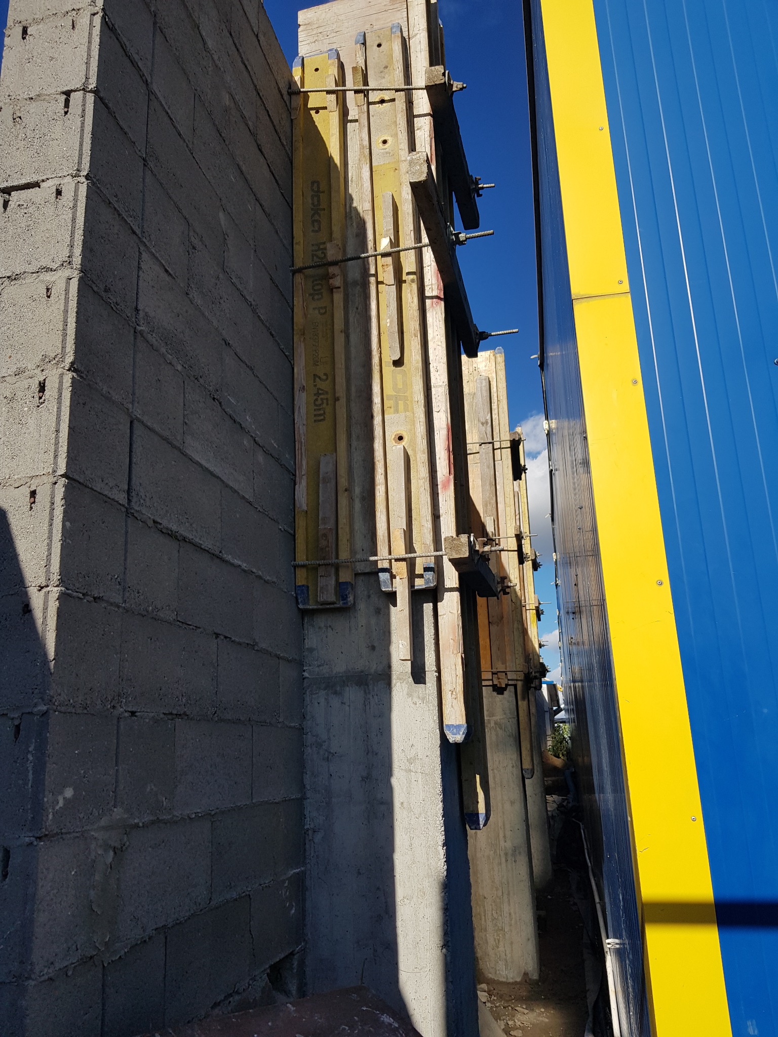 Szare bloczki fundamentowe z drewnianym szalunkiem i żółto-niebieskim panelem w tle, widok z dołu.