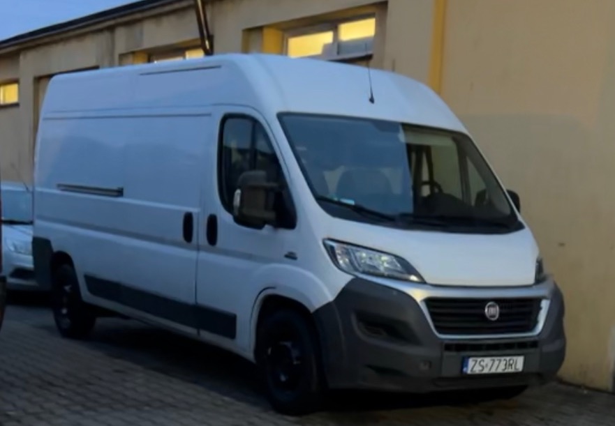 Biały samochód dostawczy Fiat Ducato z czarnymi felgami zaparkowany przed budynkiem z oknami.