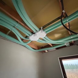 NovoInstal Damian Ogorzalek - Rozprowadzenie instalacji rekuperacji pod stropem z widoczną skrzynką rozdzielczą i przewodami wentylacyjnymi w kolorze miętowym, mocowanymi do drewnianych belek stropowych.